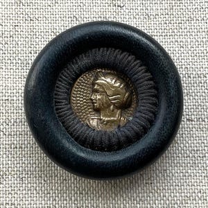 May include: A circular button featuring a smooth black outer rim and a dark, textured fabric border surrounding a central metallic gold-toned inlay. The inlay displays a relief profile of a person facing left with detailed hair and facial features.