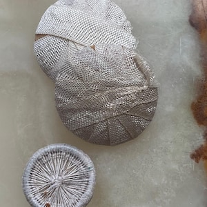 Puede incluir: Tres objetos de tela plateada tejida. Dos son redondos y en forma de moño, con un patrón texturizado en forma de rejilla. El tercero es un objeto circular más pequeño con un diseño radial. Probablemente artículos decorativos o artesanales.