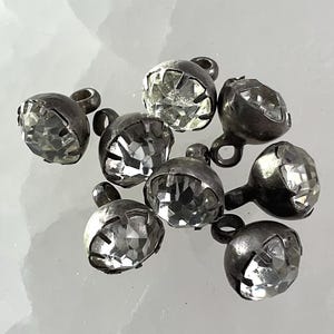 Peut inclure: Un ensemble de huit boutons métalliques ronds ornés de pierres en verre transparent à facettes, serties dans un métal argenté oxydé foncé. Chaque bouton est doté d'une petite boucle circulaire à l'arrière pour la fixation sur divers supports.