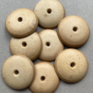 Op de afbeelding: Een verzameling van zeven ronde, lichtbeige houten knopen. Elke knoop heeft een glad, koepelvormig oppervlak met een enkel gat in het midden. De houtnerf is zichtbaar op elk stuk, wat natuurlijke variaties in textuur en tint laat zien.