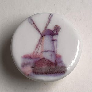 Puede incluir: Botón redondo de cerámica blanca con una imagen estilo acuarela de un molino de viento. El molino está representado en tonos morados, rosas y marrones, con las aspas extendidas. El botón tiene un acabado brillante.
