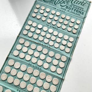 May include: A vintage light blue display card titled The Abbey Card containing 12 dozen white circular buttons. The card is divided into sections labelled 16, 18, 20, 22, and 24 lines, with rows of buttons arranged in a grid pattern on the paper surface.