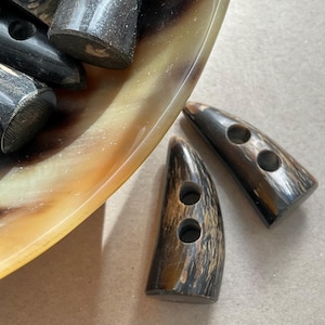 May include: A collection of dark brown and black horn toggle buttons with a smooth, polished finish. Each button features a curved, elongated shape with two circular holes drilled through the centre for attachment to garments or accessories.