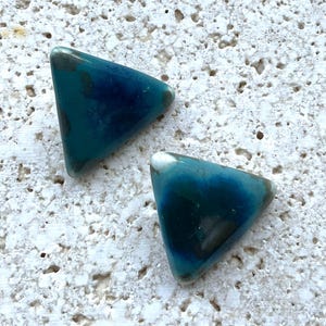 May include: Two triangular teal-colored beads with a glossy finish. The beads have a gradient of dark blue and teal, with white tips. The beads are likely for jewelry making or crafting.