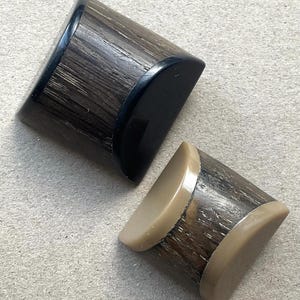 Puede incluir: Dos objetos cilíndricos con un patrón de veta de madera marrón oscuro texturizado. El objeto más grande presenta tapas negras lisas, mientras que el más pequeño tiene tapas beige lisas. Ambos artículos tienen un diseño de perfil semicircular.