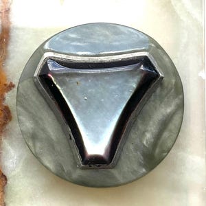 Puede incluir: Un botón redondo gris con un centro triangular de color oscuro. El botón tiene un borde metálico alrededor del triángulo. La superficie del botón tiene un aspecto veteado.