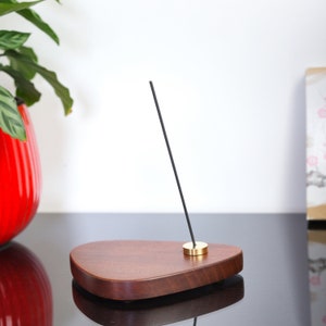 Può includere: Un porta incenso in legno marrone scuro con un top in ottone. Un bastoncino di incenso nero è in piedi nel porta incenso.