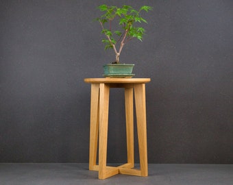 Op de afbeelding: Een kleine, lichtgekleurde houten plantenstandaard met een ronde bovenkant en vier schuine poten. Een groene bonsai in een rechthoekige pot staat erop. De plant heeft groene bladeren en een roodbruine stam. De standaard is ongeveer 61 cm hoog.