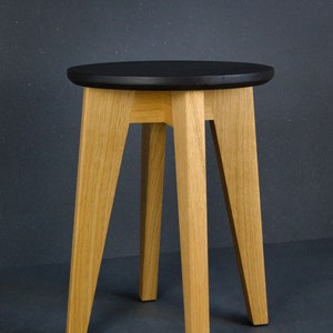 K&ouml;nnte beinhalten: Ein Holzschemel mit einer runden, schwarzen Sitzfl&auml;che und vier abgewinkelten, hellfarbigen Holzbeinen. Der Hocker hat ein schlichtes, modernes Design, das f&uuml;r verschiedene Innenr&auml;ume geeignet ist. Die H&ouml;he des Hockers betr&auml;gt ca. 46 cm.
