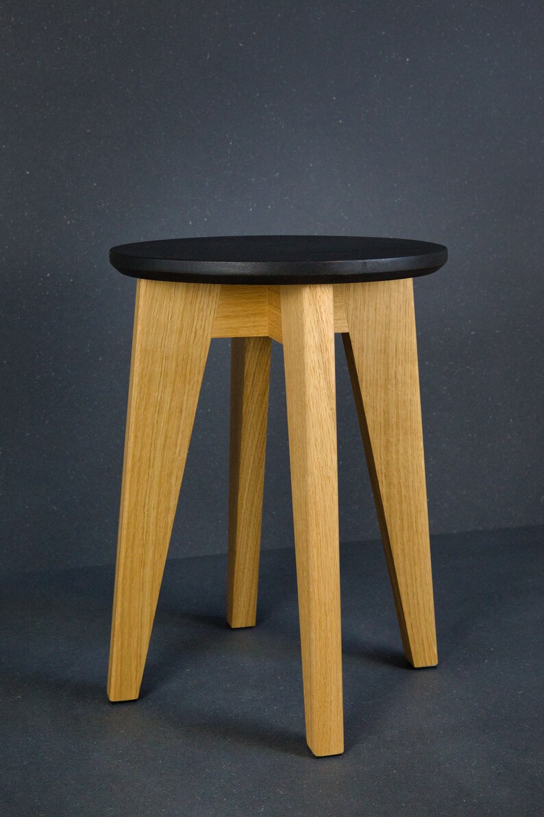 K&ouml;nnte beinhalten: Ein Holzschemel mit einer runden, schwarzen Sitzfl&auml;che und vier abgewinkelten, hellfarbigen Holzbeinen. Der Hocker hat ein schlichtes, modernes Design, das f&uuml;r verschiedene Innenr&auml;ume geeignet ist. Die H&ouml;he des Hockers betr&auml;gt ca. 46 cm.