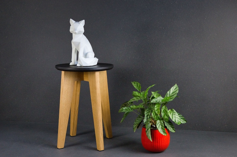 K&ouml;nnte beinhalten: Eine wei&szlig;e, geometrische Fuchsskulptur sitzt auf einem kleinen, runden, schwarzen Tisch mit Beinen aus Naturholz. Rechts steht ein roter Topf mit einer gr&uuml;nen Pflanze. Der Hintergrund ist dunkelgrau.