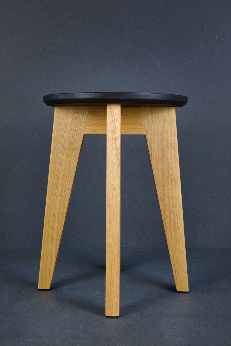 K&ouml;nnte beinhalten: Ein Holzschemel mit einer runden, schwarzen Sitzfl&auml;che und drei hellen, abgewinkelten Beinen. Der Hocker ist aus Naturholz mit sichtbarer Maserung gefertigt und steht auf einer dunkelgrauen Oberfl&auml;che. Das Design ist minimalistisch und modern.