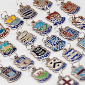 May include: A collection of small, shield-shaped metal charms featuring colorful enamel designs and various town names. Each charm includes a small silver-toned jump ring at the top for attachment, displaying distinct heraldic-style emblems and text.