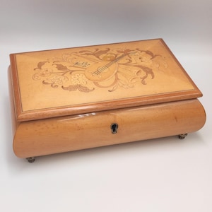May include: A light-coloured wooden music box with an inlaid design on the lid. The lid features a musical instrument and floral patterns. The box has a curved body, a keyhole, and small feet.