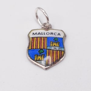 May include: A silver-toned pendant with a shield-shaped design. The shield features a colorful emblem with the word "MALLORCA" at the top. The emblem includes a blue background with a castle and palm trees, and vertical stripes.
