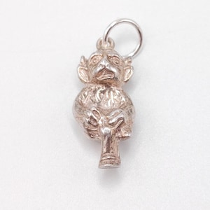 Peut inclure: Un charm argenté en forme de singe assis. Le charme présente un visage détaillé, de grandes oreilles et un corps texturé. Une petite boucle est attachée en haut pour l'accrocher. Le charme est probablement en argent sterling et est posé sur un fond blanc.