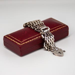 Puede incluir: Una pulsera de plata con un diseño de cadena tejida de múltiples eslabones y un dije de candado en forma de corazón. La pulsera descansa sobre un joyero rectangular de color rojo oscuro con un borde decorativo dorado.