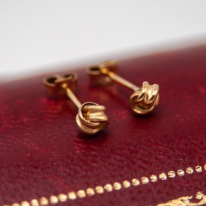 May include: A pair of gold stud earrings featuring a small, intricate knot design at the front. Each earring has a thin post extending from the back of the knot, designed to be secured with a matching gold earring back component.