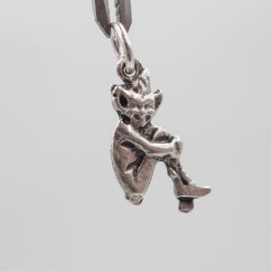 Puede incluir: Un amuleto de plata en forma de hada sentada con alas. El hada tiene una cara detallada y lleva un vestido.