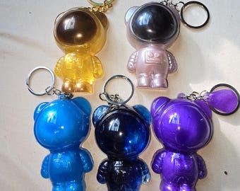May include: Six astronaut bear keychains in different colors: gold, silver, light pink, light blue, dark blue, and purple. Each keychain features a bear wearing an astronaut helmet.