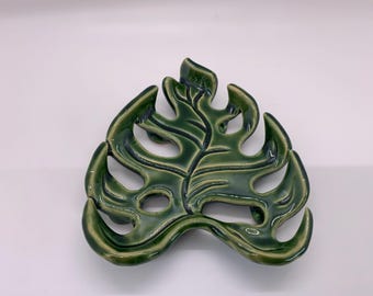 May include: A glossy, dark green ceramic trivet shaped like a monstera leaf. The piece features deep, carved veins and open cutouts between the leaf segments, with a smooth, reflective finish that highlights the organic, curved edges of the design.
