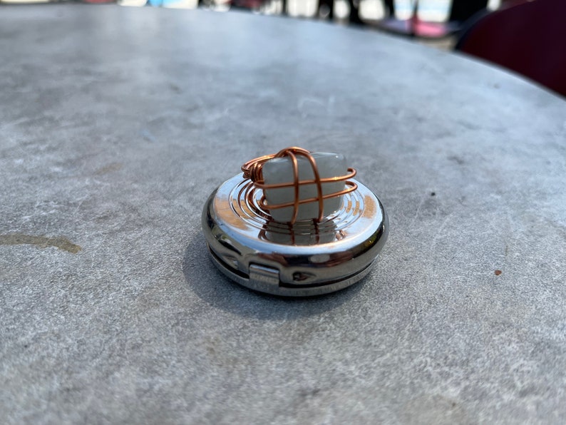 Puede incluir: Un porta anillos de plata con un anillo de piedra blanca envuelto en alambre de cobre.