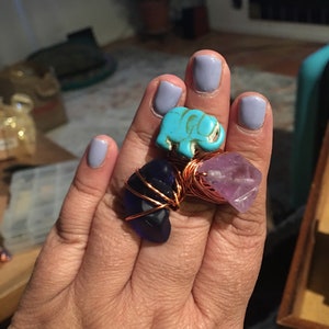 Puede incluir: Tres anillos con diferentes piedras y alambre de cobre envuelto. El primer anillo presenta un amuleto de elefante turquesa. El segundo anillo tiene una piedra de vidrio azul oscuro. El tercer anillo tiene una piedra de amatista p&uacute;rpura.