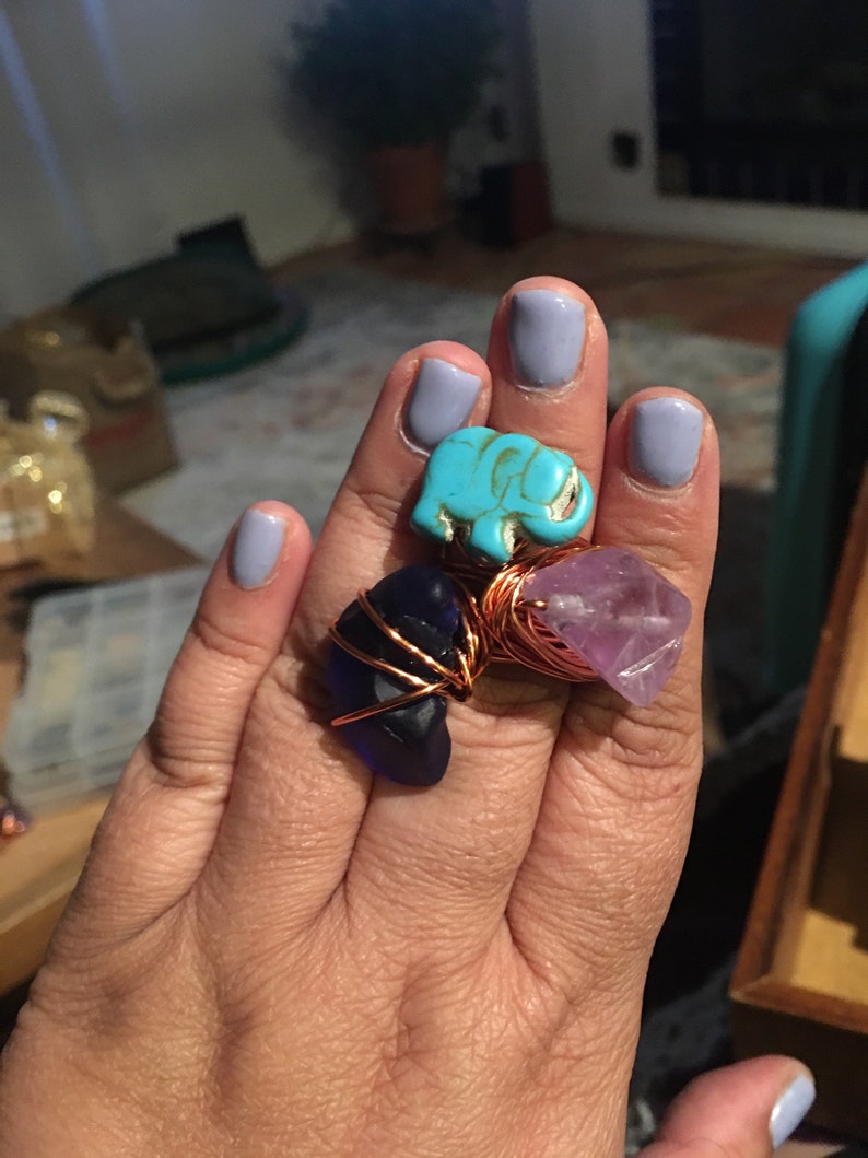 Puede incluir: Tres anillos con diferentes piedras y alambre de cobre envuelto. El primer anillo presenta un amuleto de elefante turquesa. El segundo anillo tiene una piedra de vidrio azul oscuro. El tercer anillo tiene una piedra de amatista p&uacute;rpura.
