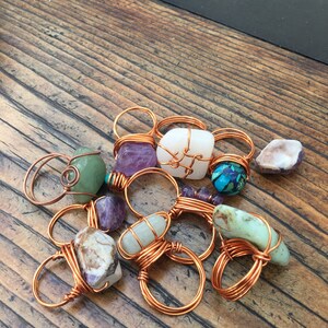 Puede incluir: Una colecci&oacute;n de anillos envueltos en alambre de cobre con varias piedras semipreciosas, incluyendo amatista, turquesa y &aacute;gata. Los anillos se muestran sobre una superficie de madera.
