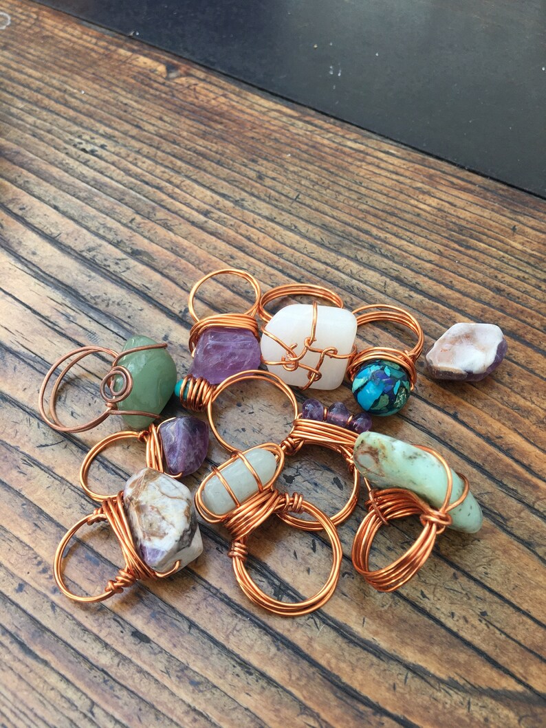 Puede incluir: Una colecci&oacute;n de anillos envueltos en alambre de cobre con varias piedras semipreciosas, incluyendo amatista, turquesa y &aacute;gata. Los anillos se muestran sobre una superficie de madera.