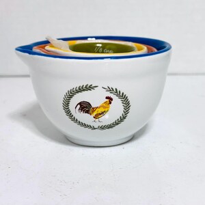 Può includere: Una ciotola in ceramica bianca con bordo blu e un motivo di gallo colorato. All'interno sono presenti misurini ad incastro gialli, arancioni e verdi. Il misurino più piccolo è etichettato "1/8 cup".