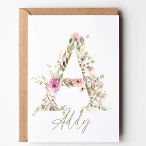 May include: A white greeting card featuring a large letter A formed by watercolor floral illustrations in shades of pink, green, and brown. Below the letter, the name Addy is written in a grey cursive script. The card is tucked into a brown envelope.