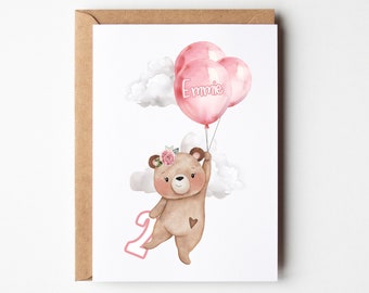 May include: A greeting card featuring a brown bear with a pink floral crown and a heart patch, holding three pink balloons. One balloon displays the name Emmie in white text. A pink number two is positioned beside the bear, with white clouds.