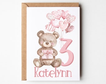 May include: A white greeting card featuring a watercolor illustration of a brown teddy bear holding a pink gift box. The bear is next to a large pink number 3 and a cluster of pink heart-shaped balloons. The name Katelynn is printed in pink below.