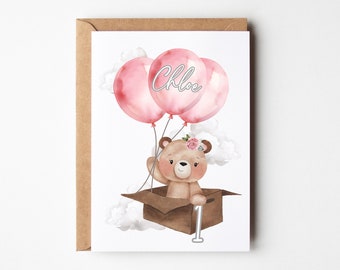 May include: A greeting card featuring a watercolor illustration of a brown teddy bear sitting in a cardboard box, holding three pink balloons. The center balloon has the name Chloe written on it. A white number one is next to the box.