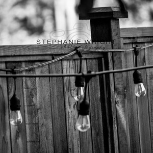 May include: Black and white photograph of a wooden fence with string lights. The lights are hanging from a wire and have a classic bulb shape. The fence is made of vertical wooden planks. The image includes the text "STEPHANIE WRIGHT photography".
