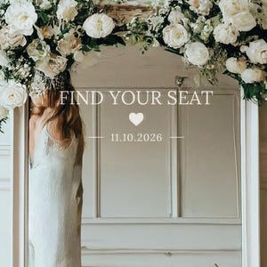 Op de afbeelding: Een grote wit omkaderde spiegel met de tekst "FIND YOUR SEAT" en de datum 11.10.2026. De spiegel is versierd met witte bloemen en groen bovenaan. De weerspiegeling toont een bank en een persoon in een witte jurk.