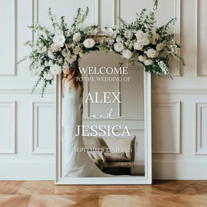 Puede incluir: Un espejo rectangular grande con marco blanco, adornado con arreglos florales blancos. El espejo muestra el texto "WELCOME TO THE WEDDING OF ALEX and JESSICA SEPTEMBER 22ND 2025" en letras blancas, ideal para una boda.