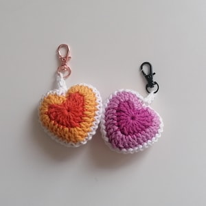 Può includere: Due portachiavi a forma di cuore fatti a mano all'uncinetto. Un cuore è arancione e giallo, l'altro è rosa e viola. Entrambi hanno bordi bianchi e chiusure in metallo. Un accessorio affascinante e colorato.
