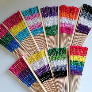 Könnte beinhalten: Eine Sammlung farbenfroher Handfächer mit Holzgriffen. Jeder Fächer hat eine gehäkelte Oberseite in einem anderen Regenbogen- oder Pride-Flaggen-Farbschema, darunter Rosa, Blau, Gelb, Grün, Lila und Schwarz.