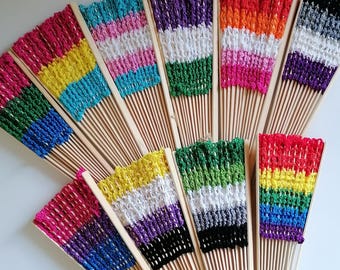 Puede incluir: Una colección de nueve abanicos con varillas de madera y partes superiores de tela tejida a ganchillo con diversos patrones de rayas multicolores. Cada abanico muestra una combinación única de colores de hilo vibrantes en bandas horizontales.