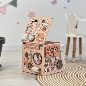 May include: A light pink wooden activity cube with various interactive elements, including gears, switches, and shapes. The cube is open, revealing additional components. The background features a plant, a rug, and a child's room decor.