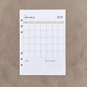 May include: A white planner insert page for January 2027 featuring a monthly calendar grid with a beige header row. The page includes six punched holes along the left side, with sections labelled Notes and Top Priorities at the bottom.