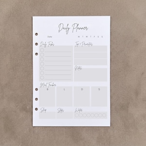 May include: A white daily planner insert with six binder holes on the left side. The page features sections for Daily Tasks with checkboxes, Top 3 Priorities, Notes, a Meal Tracker for B, L, D, S, and trackers for Sleep, Steps, and Water intake.