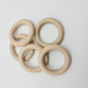 Könnte beinhalten: Fünf unfertige Holzringe, jeweils etwa 7,6 cm im Durchmesser, liegen auf einem weißen Hintergrund. Die Ringe bestehen aus hellem Holz und zeigen die natürliche Maserung. Diese Holzringe eignen sich für Bastelprojekte.