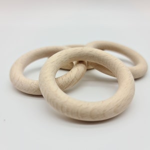 Könnte beinhalten: Drei unfertige Holzringe, wahrscheinlich für Bastelarbeiten oder Babyspielzeug, liegen auf einer weißen Oberfläche. Die hellfarbigen Ringe zeigen die natürliche Holzmaserung. Die Anordnung deutet auf einen spielerischen oder pädagogischen Zweck hin.