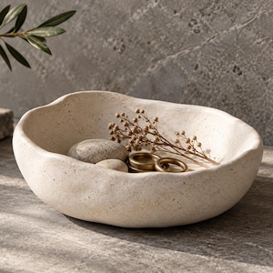 May include: A shallow, off-white ceramic bowl with an irregular, organic rim and a speckled texture. Inside the bowl are two smooth gray stones, two small gold-toned metal rings, and a delicate, dried brown plant sprig with tiny seed pods.