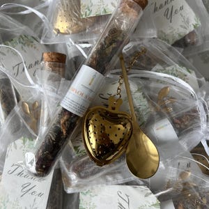 Pode incluir: Uma coleção de itens relacionados ao chá, incluindo um tubo de vidro cheio de chá solto, um infusor de chá dourado em forma de coração e uma pequena colher dourada. Cada item é embalado em um saco branco transparente com uma etiqueta "Obrigado", sugerindo um presente.