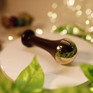 May include: A dark brown facial massage tool with a gold-coloured, rounded end, resting on a white surface. The tool has a cylindrical shape with a bulbous end. The background includes greenery and soft, out-of-focus lights.