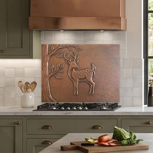 May include: A rectangular copper backsplash panel featuring a raised relief design of a deer standing next to a tree. The metallic panel is installed behind a black gas stovetop, set against a kitchen wall with olive green cabinets and white tiles.